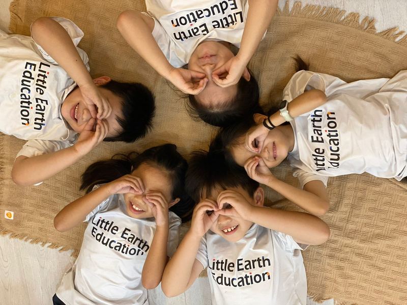 Children in Little Earth T-shirts lie in a circle with their heads together, making goggles with their fingers
