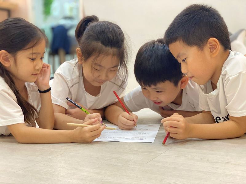 Four children in Little Earth T-shirts lie on the floor around one worksheet, pencils in hand, smiling as they work it out together