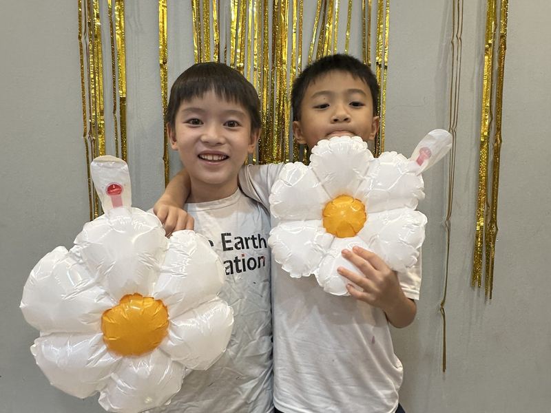 Two boys, one in a Little Earth T-shirt, hold big flower-shaped balloons in front of a gold tinsel curtain, one grinning at the camera