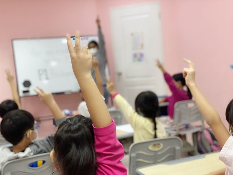 Children in a classroom raise their hands to answer, seen from behind