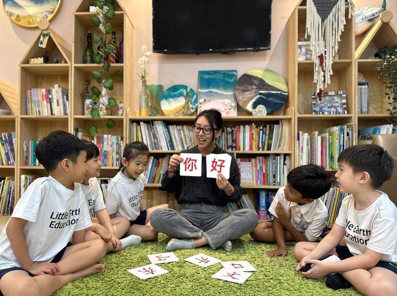A teacher holds up Chinese character cards reading 你好 to children sitting on the rug in the reading corner