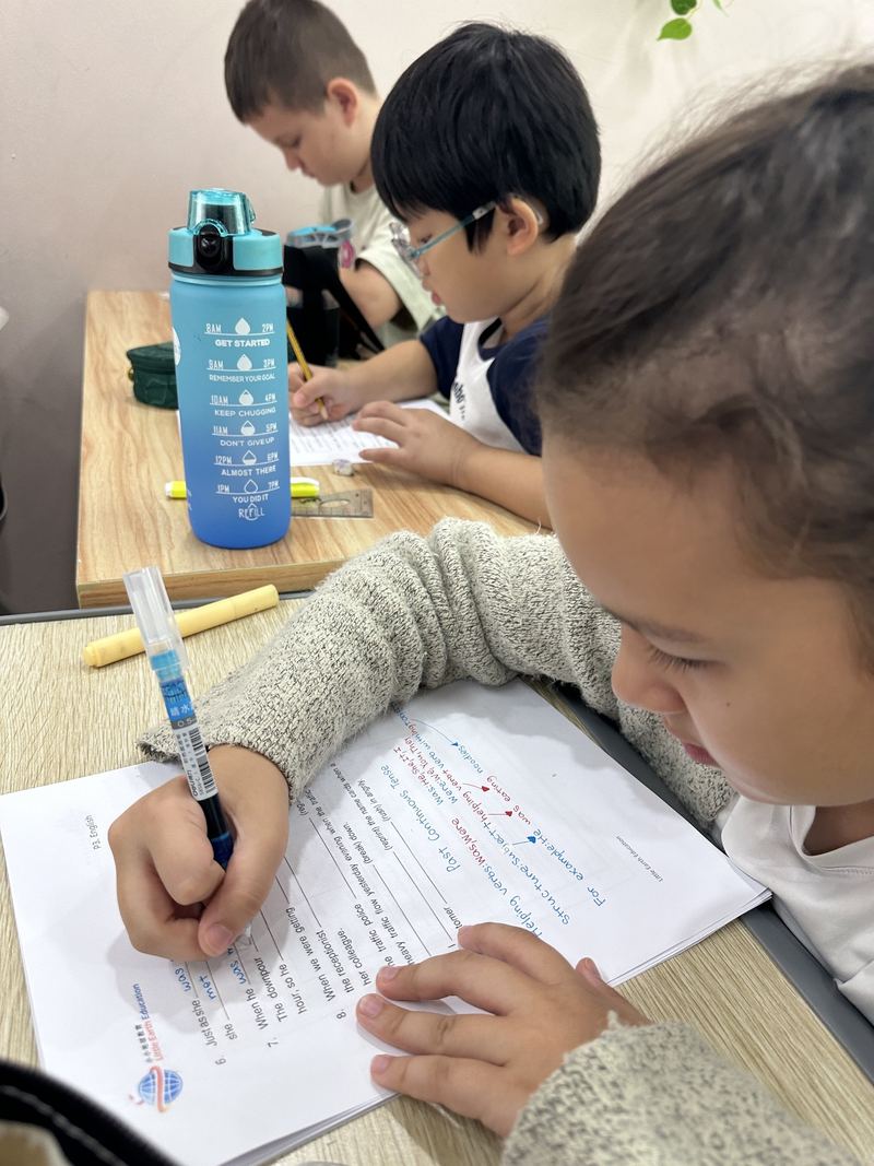 A child writes answers on an English worksheet at a desk, a water bottle beside the book