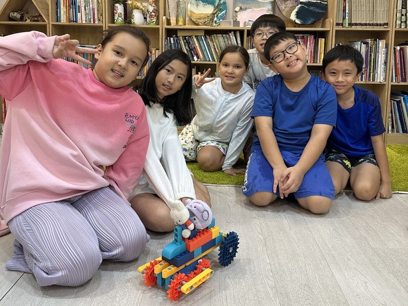 Six children kneel by the bookshelves, smiling and waving, with the robot they built
