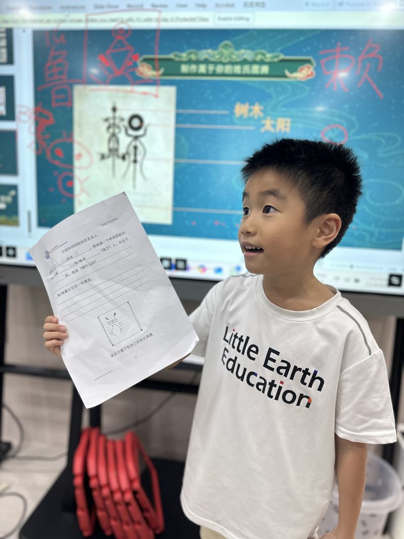 A boy in a Little Earth T-shirt holds up his Chinese worksheet in front of the class screen